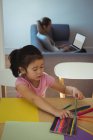 Chica seleccionando el lápiz de color mientras que la madre usando el ordenador portátil en el fondo en casa - foto de stock