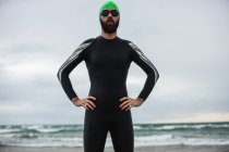 Retrato de atleta en traje mojado de pie con las manos en la cintura en la playa - foto de stock