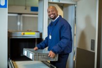 Портрет улыбающегося сотрудника службы безопасности аэропорта с ящиком рядом с конвейерной лентой в терминале аэропорта — стоковое фото