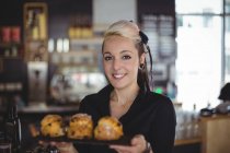 Портрет офіціантки тримає лоток кексів за лічильником в кафе — стокове фото