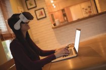 Женщина, использующая гарнитуру виртуальной реальности во время ввода на ноутбуке дома — стоковое фото