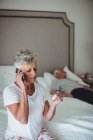 Занепокоєна старша жінка сидить у спальні, тримає ліки і розмовляє на мобільному телефоні — стокове фото