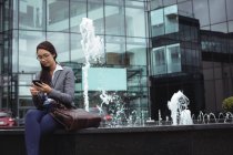 Бізнес-леді сидить біля фонтану і використовує мобільний телефон за межами офісної будівлі — стокове фото