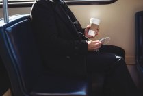 Средняя секция бизнесмена, держащего одноразовый кофейный стаканчик и пользующегося мобильным телефоном в поезде — стоковое фото