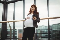 Embarazada mujer de negocios utilizando el teléfono móvil cerca del pasillo en la oficina - foto de stock