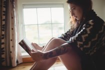Женщина сидит у окна и читает книгу дома — стоковое фото