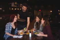 Amigos felizes desfrutando de comida no bar enquanto garçom serve comida — Fotografia de Stock