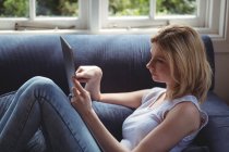 Belle femme assise sur un canapé et utilisant une tablette numérique dans le salon à la maison — Photo de stock