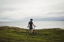 Atleta de pé com sua bicicleta na grama perto do mar — Fotografia de Stock
