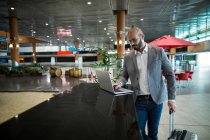Бізнесмен стоїть з сумкою на візку, використовуючи ноутбук у зоні очікування в терміналі аеропорту — стокове фото