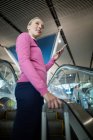 Comutador feminino com bagagem usando telefone celular na escada rolante no aeroporto — Fotografia de Stock