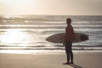 Uomo che trasporta tavola da surf in piedi sulla spiaggia al tramonto — Foto stock