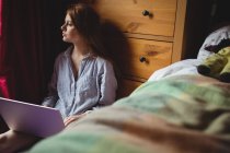 Задумчивая женщина использует ноутбук в спальне дома — стоковое фото