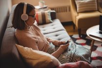 Woman sitting on sofa listening to music on mobile phone in living room at home — Stock Photo