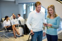 Couple standing with luggage holding smartphone and boarding pass in waiting area — Stock Photo