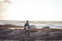 Homem carregando uma prancha de surf caminhando em direção ao mar ao anoitecer — Fotografia de Stock