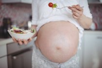Sección media de la mujer embarazada que tiene ensalada en la cocina en casa - foto de stock
