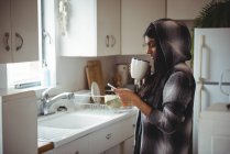 Жінка стоїть на кухні за допомогою мобільного телефону вдома — стокове фото