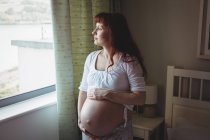 Задумчивая беременная женщина смотрит в окно в спальне дома — стоковое фото
