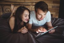 Couple couché sur le lit en utilisant une tablette numérique dans la chambre à coucher à la maison — Photo de stock