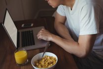 Homem usando telefone celular e laptop enquanto toma café da manhã no quarto em casa — Fotografia de Stock