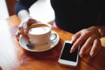 Sección media de la mujer usando el teléfono móvil mientras toma una taza de café en la cafetería - foto de stock
