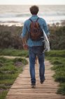 Visão traseira de um homem carregando uma prancha andando no calçadão — Fotografia de Stock