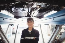 Женщина-механик, стоящая с скрещенными руками под машиной в ремонтном гараже — стоковое фото