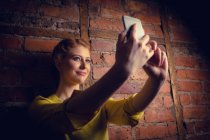 Donna in piedi contro muro di mattoni e prendere un selfie sul suo telefono cellulare — Foto stock