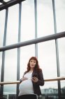 Беременная деловая женщина, использующая мобильный телефон рядом с коридором в офисе — стоковое фото