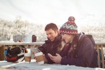 Щаслива пара лижників сидить за столом, використовуючи мобільний телефон та цифровий планшет у гірськолижному курорті — стокове фото