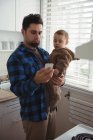 Отец использует мобильный телефон, держа ребенка дома на кухне — стоковое фото