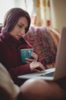 Красива жінка, що має каву, використовуючи ноутбук на дивані вдома — стокове фото