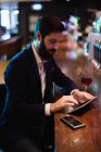 Empresario que utiliza tableta digital con copa de vino y teléfono móvil en el mostrador en el bar - foto de stock