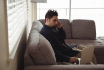 Мужчина разговаривает по мобильному телефону, используя ноутбук на диване дома — стоковое фото