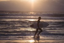 Silhouette of a man carrying surfboard walking on beach at dusk — Stock Photo