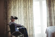 Пенсионер сидит на инвалидной коляске и пользуется гарнитурой виртуальной реальности в спальне дома — стоковое фото