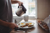 Мужчина держит тарелку с завтраком, наливая горячую воду в кружку на кухне. — стоковое фото