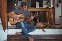 Мужчина играет на гитаре дома, собака лежит рядом с ним — стоковое фото