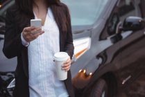 Женщина, стоящая рядом с автомобилем и использующая мобильный телефон на зарядной станции электромобиля — стоковое фото
