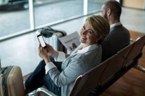 Портрет усміхненої бізнес-леді з мобільним телефоном, що сидить у зоні очікування в терміналі аеропорту — стокове фото
