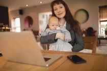 Happy mother sitting at table and holding baby at home — Stock Photo