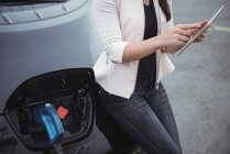 Mittelteil der Frau nutzt digitales Tablet beim Laden von Elektroautos auf der Straße — Stockfoto