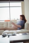 Вагітна жінка цифровий планшет, розслабляючись на дивані у вітальні вдома — стокове фото