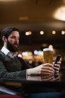 Homme regardant téléphone portable tout en prenant un verre de bière dans le bar — Photo de stock