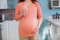 Mittlerer Abschnitt der schwangeren Frau hält Glas Wasser in der Küche zu Hause — Stockfoto