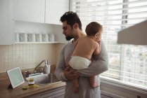 Vater schaut auf digitales Tablet, während er seinen kleinen Sohn in der Küche hält — Stockfoto