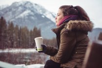 Vista lateral de la mujer sentada en la orilla del río sosteniendo la taza de café en invierno - foto de stock