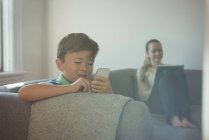 Сын использует мобильный телефон и его мать в фоновом режиме дома — стоковое фото