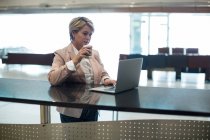 Empresaria que usa laptop mientras toma café en la sala de espera en la terminal del aeropuerto - foto de stock
